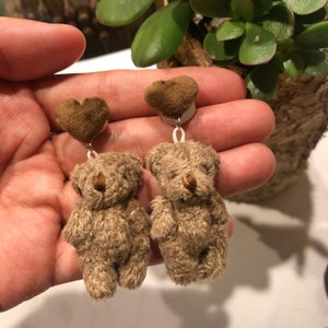 May include: A pair of brown teddy bear dangle earrings with brown heart-shaped charms. The earrings are made of soft, fuzzy fabric and have a delicate, whimsical design.
