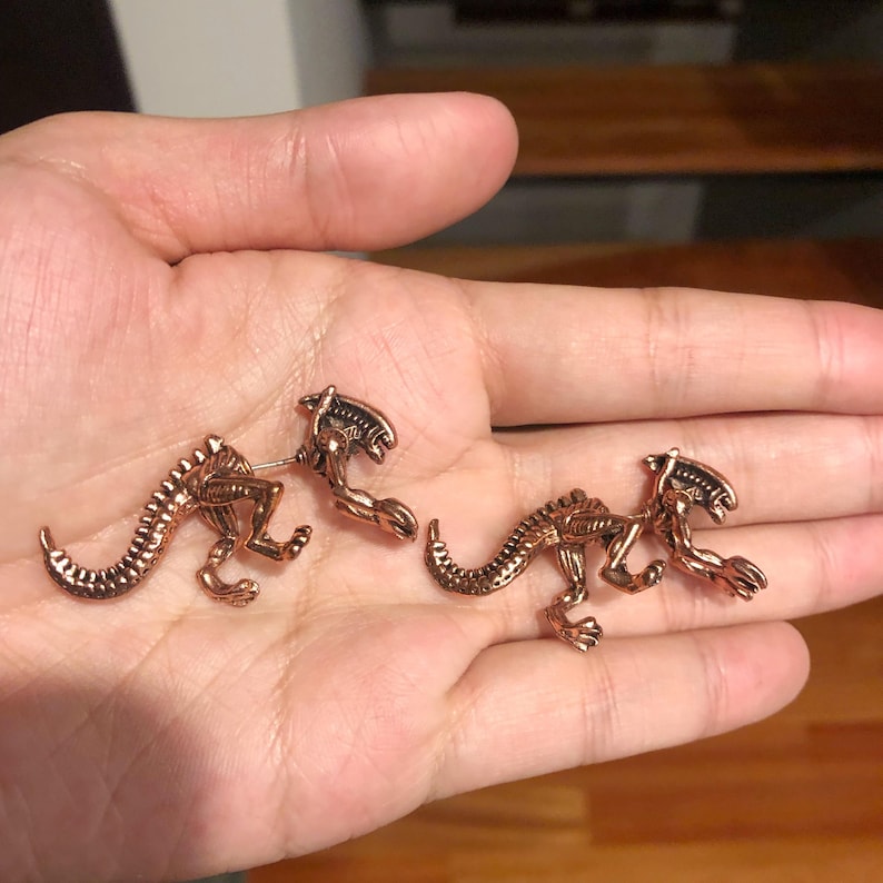 May include: A pair of bronze-colored alien creature earrings. The earrings are in the shape of a small alien creature with a long tail and a head with a large mouth.
