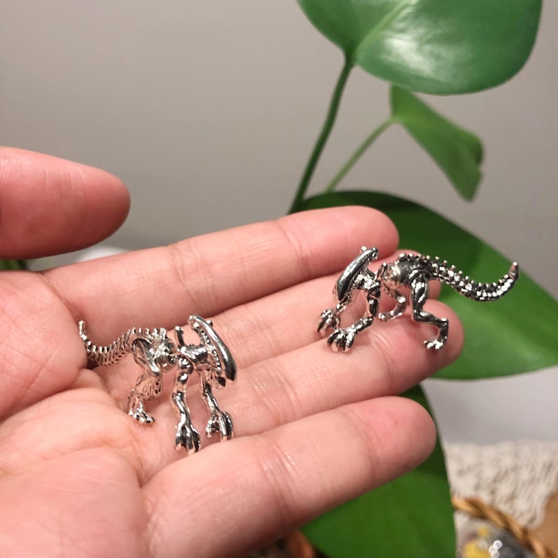 May include: Two silver alien creature earrings. The earrings are in the shape of a small alien with a long tail. The earrings are shown on a hand.