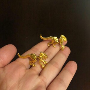 May include: A pair of gold-colored alien-shaped earrings. The earrings are small and detailed, with a textured surface.