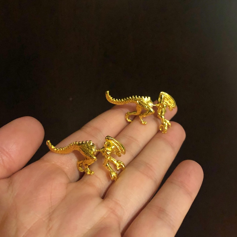 May include: A pair of gold-colored alien-shaped earrings. The earrings are small and detailed, with a textured surface.