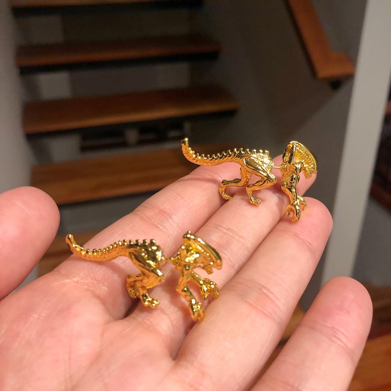 May include: Two gold-colored alien creature earrings. The earrings are in the shape of a small alien creature with a long tail and a raised head. The earrings are shown on a hand.