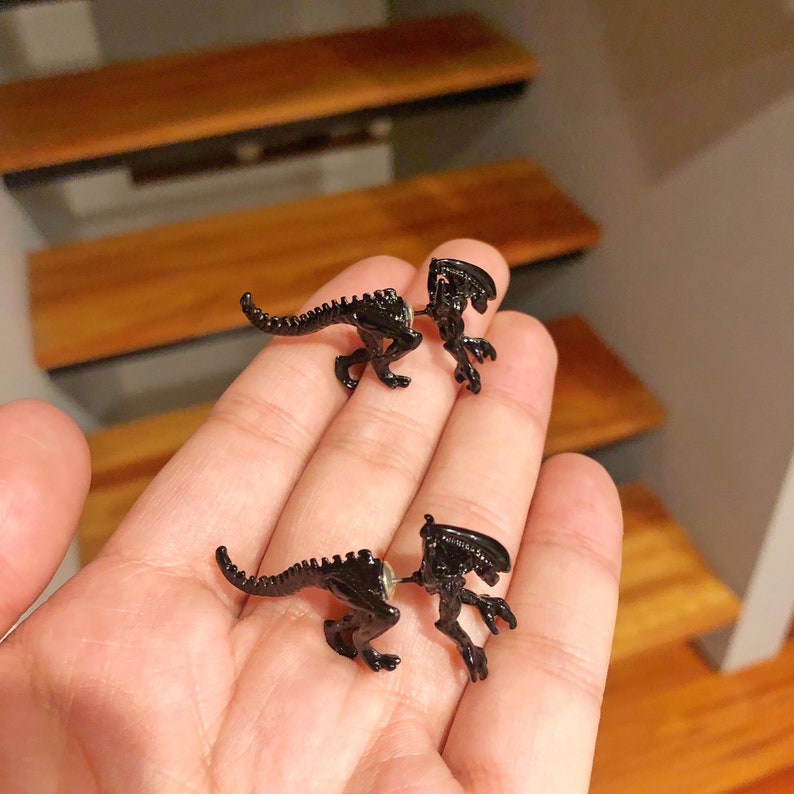 May include: A pair of black alien-shaped earrings. The earrings are made of metal and have a detailed design. The earrings are shown on a person's hand.