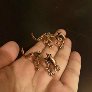 May include: A pair of gold-toned alien-shaped earrings. The earrings are detailed with a skeletal design and have a textured finish.