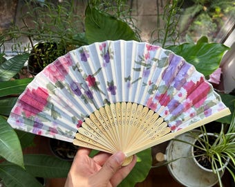 May include: A white folding fan with a blue and pink floral pattern. The fan has a wooden handle with a decorative design.