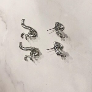 May include: A pair of silver alien-shaped earrings. The earrings are in the shape of an alien with a long tail and a large head.