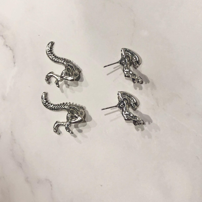 May include: A pair of silver alien-shaped earrings. The earrings are in the shape of an alien with a long tail and a large head.