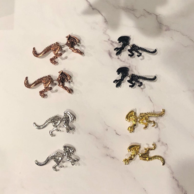 May include: Eight pairs of alien-shaped earrings in silver, gold, and black metal. The earrings are all in the same style, but with different finishes.