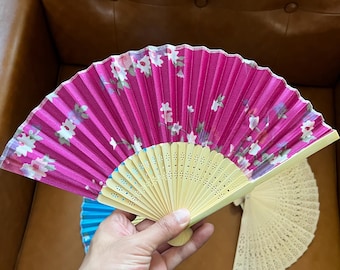 May include: Two hand fans, one pink with white floral print and one blue with white floral print. Both fans have a wooden frame with intricate cutouts.