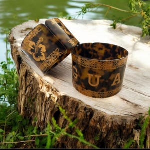 May include: Three brown and gold bangles with a tortoiseshell pattern are displayed on a tree stump. One bangle features a hibiscus flower design, while another displays the letters "GUA". The bangles are surrounded by greenery and water.