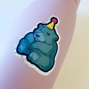 May include: A light purple water bottle with a holographic sticker of a cartoon bear wearing a party hat. The bear is blue and green and has a grumpy expression.