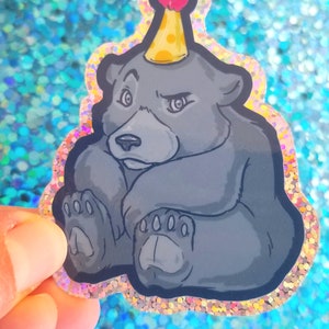May include: A black bear cub wearing a yellow and red party hat with a white pom-pom. The bear is sitting with its paws crossed and has a grumpy expression. The sticker is on a glittery blue background.