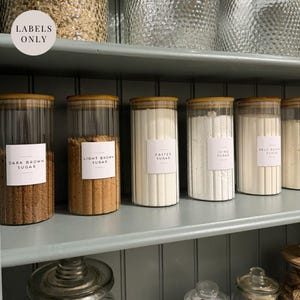 May include: Six glass jars with wooden lids and white labels on a grey shelf. The labels read "Dark Brown Sugar", "Light Brown Sugar", "Caster Sugar", "Icing Sugar", and "Self Raising Flour".