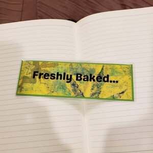 May include: A lined notebook with a green and yellow bookmark that says "Freshly Baked..."