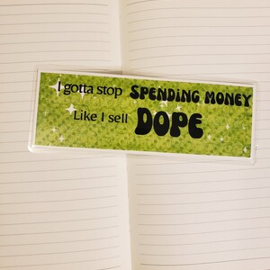 May include: A green bookmark with a white background and glitter. The bookmark has the text "I gotta stop spending money like I sell dope" in black text.