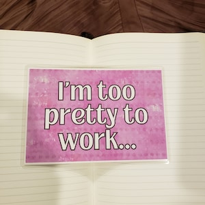 May include: An open notebook with lined pages, featuring a pink card with the text "I'm too pretty to work..." in white. The card is in a clear plastic sleeve. The background is a brown, textured fabric.
