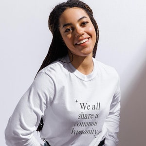 May include: A woman wearing a white long-sleeved shirt with the text "We all share a common humanity" printed on the front. She is wearing blue jeans and has her hands in her pockets.