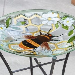 May include: An oval glass bird bath with a bee and honeycomb design. The dish features a painted bee with yellow and black stripes, white flowers, and green leaves. The bird bath is supported by a black metal stand.