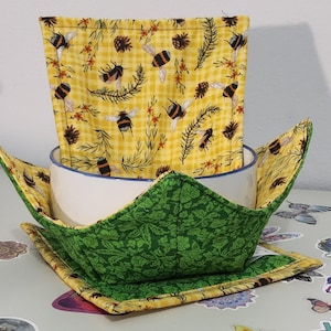 May include: A white bowl with a blue rim sits inside a green and yellow fabric bowl cozy. The cozy has a green leaf pattern on the outside and a yellow bee and pinecone pattern on the inside. A matching yellow bee-themed square fabric is inside the bowl.