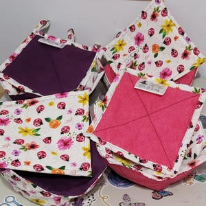 May include: A collection of handmade fabric bowl cozies and pot holders. The exterior fabric features a white background with a floral and ladybug pattern. The interior fabric is solid purple or pink. Each item has a tag.