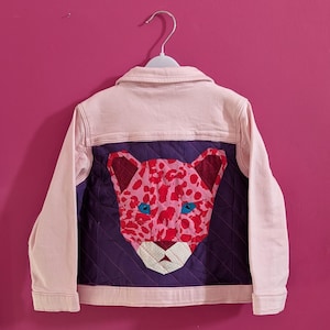 May include: A light pink denim jacket with a purple quilted back panel. The back features a colorful leopard face applique with pink spots, red accents, and blue eyes. The jacket is hanging on a white hanger against a pink wall.