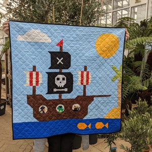 May include: A blue and brown quilted baby blanket with a pirate ship design. The ship has a skull and crossbones flag, a red flag, and a white cloud above it. There are two yellow fish and a palm tree on the blanket.