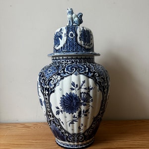 Puede incluir: Un jarrón de cerámica azul y blanco con tapa. El jarrón tiene un diseño floral y una pequeña figura de animal en la parte superior de la tapa.