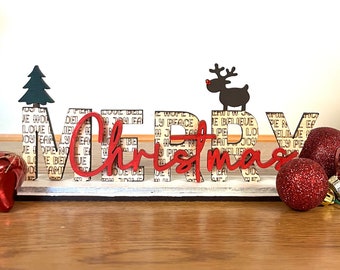 Può includere: Un cartello di legno con le parole "Merry Christmas" in lettere rosse. Il cartello è decorato con un piccolo albero verde, una renna nera con corna rosse e palline rosse e argentate.
