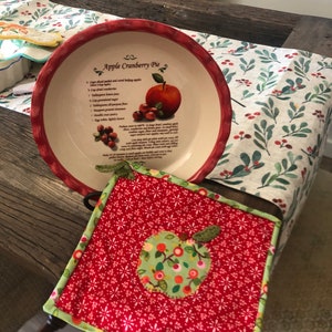 Peut inclure: Un dessous de plat rouge et vert sur le thème de la pomme avec un fond blanc et une bordure rouge. Le dessous de plat a un motif de pomme verte au centre avec un fond blanc et des détails rouges et verts. Le dessous de plat est sur une table en bois avec un chemin de table blanc et vert à motif de feuilles et de baies.