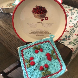 Könnte beinhalten: Ein weißer Keramik-Kuchenteller mit rotem Rand und einem darauf gedruckten Rezept für Kirschkuchen. Das Rezept enthält Zutaten wie Butter, Zucker, Kirschen und Tapioka. Der Teller steht auf einem Tisch mit einer rot-weiß karierten Tischdecke. Im Vordergrund befindet sich ein gehäkeltes Topflappen mit Kirschen.