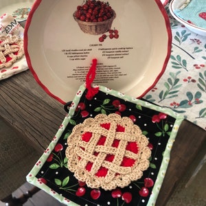 Könnte beinhalten: Eine weiße Keramik-Tortenform mit rotem Rand und einem aufgedruckten Rezept für Kirschkuchen. Das Rezept enthält Zutaten wie Butter, Kirschen und Zucker. Die Form steht auf einem Holztisch neben einem schwarz-weißen Kirsch-Themen-Topflappen mit einem gehäkelten Kirschkuchen-Design.