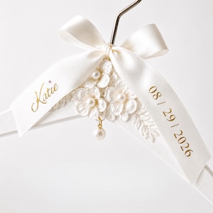 May include: A white clothes hanger adorned with a cream satin ribbon tied in a bow. The ribbon features the name Katie and the date 08 / 29 / 2026 printed in gold. Three white fabric flowers with pearl centers and a dangling pearl charm are attached.