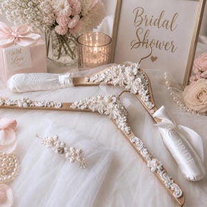 May include: Two wooden bridal hangers with white floral detailing and pearl accents. One hanger has a white satin ribbon with "Bride" embroidered. A framed sign reads "Bridal Shower". The scene includes a gift box, flowers, and a candle.