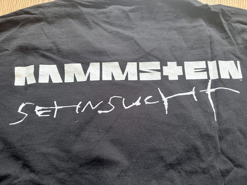 May include: Black t-shirt with the band name "RAMMSTEIN" in large, white, block letters across the top. Below, in a handwritten style, is the phrase "SEHNSUCHT". The shirt is slightly wrinkled.