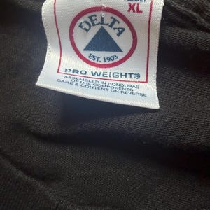 May include: Close-up of a black t-shirt tag. The tag is white with red and blue text and a logo that reads "DELTA EST. 1903". The tag also says "PRO WEIGHT" and "ADULT XL".