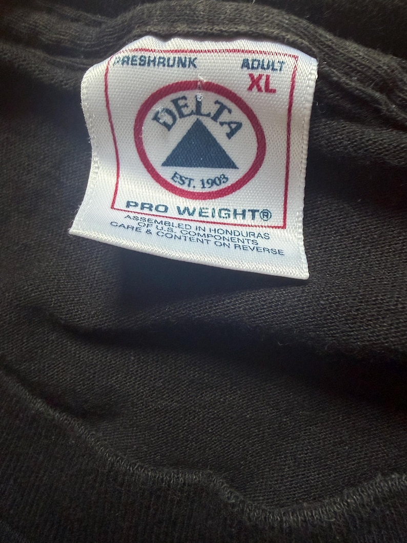 May include: Close-up of a black t-shirt tag. The tag is white with red and blue text and a logo that reads "DELTA EST. 1903". The tag also says "PRO WEIGHT" and "ADULT XL".