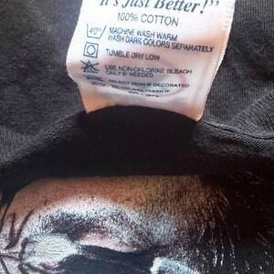 May include: Close-up of a black t-shirt with a white tag. The tag reads "It's Just Better!" and includes washing instructions: machine wash warm, tumble dry low, and use non-chlorine bleach if needed. The shirt is 100% cotton.