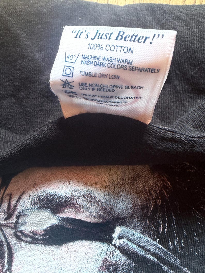 May include: Close-up of a black t-shirt with a white tag. The tag reads "It's Just Better!" and includes washing instructions: machine wash warm, tumble dry low, and use non-chlorine bleach if needed. The shirt is 100% cotton.