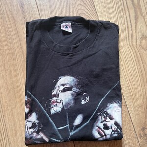 May include: Black t-shirt featuring a graphic design of three faces with metal restraints. The design is in shades of gray and red. The shirt is folded on a wooden surface.