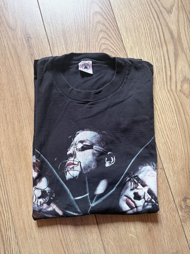 May include: Black t-shirt featuring a graphic design of three faces with metal restraints. The design is in shades of gray and red. The shirt is folded on a wooden surface.