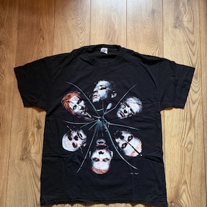 May include: Black t-shirt featuring a graphic of six faces arranged in a circular pattern, with a spiderweb design. The faces have a pale complexion and are surrounded by a dark background. The shirt is made of a soft material.