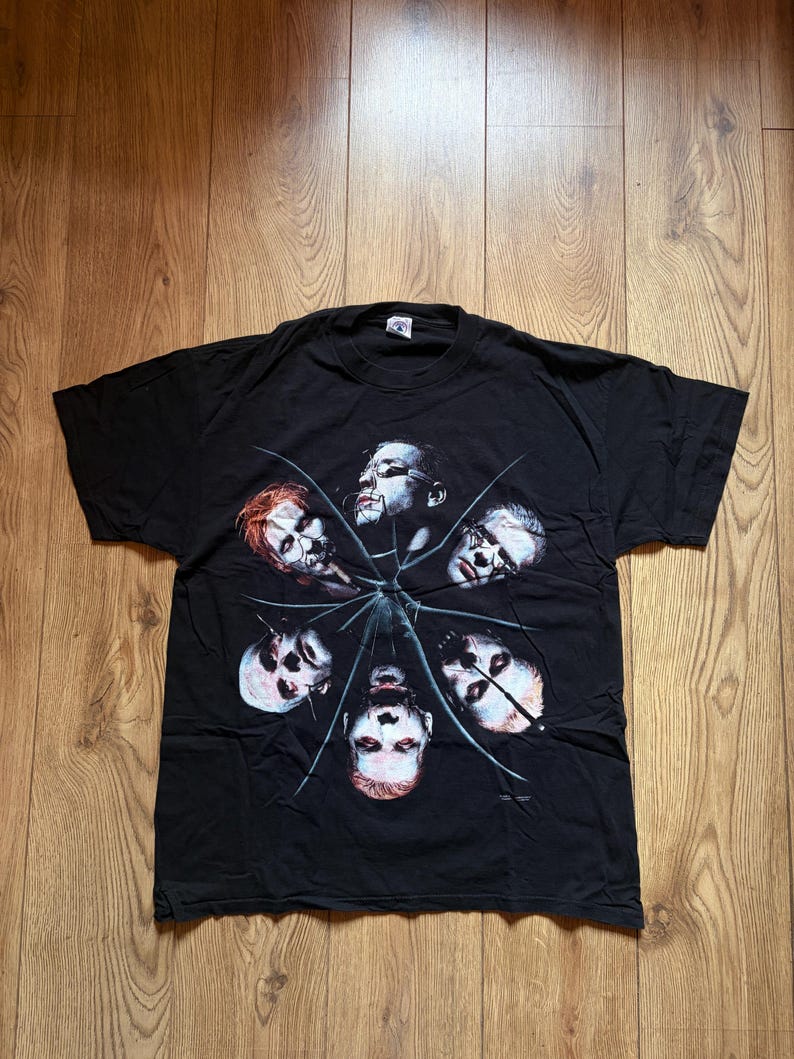 May include: Black t-shirt featuring a graphic of six faces arranged in a circular pattern, with a spiderweb design. The faces have a pale complexion and are surrounded by a dark background. The shirt is made of a soft material.