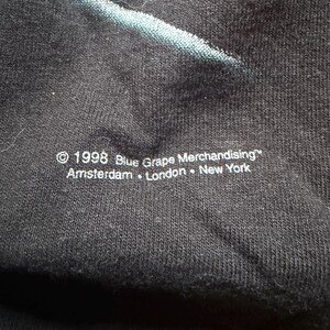 May include: Close-up of a black fabric with a faded blue design. The text "&copy; 1998 Blue Grape Merchandising&trade; Amsterdam &bull; London &bull; New York" is printed in white at the bottom. The fabric has a textured appearance.