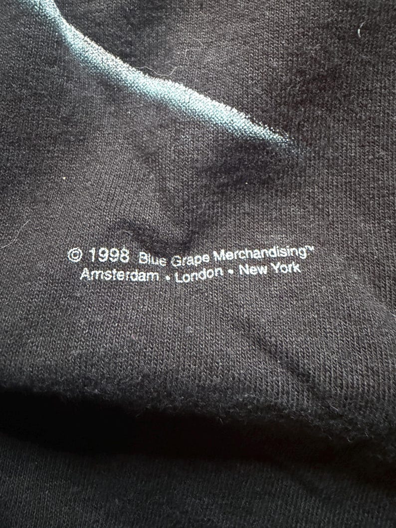 May include: Close-up of a black fabric with a faded blue design. The text "&copy; 1998 Blue Grape Merchandising&trade; Amsterdam &bull; London &bull; New York" is printed in white at the bottom. The fabric has a textured appearance.
