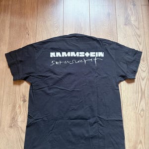 May include: Black t-shirt with the band name "RAMMSTEIN" in white block letters above the word "Sehnsucht" in a stylized white font. The shirt is laid flat on a wooden surface.