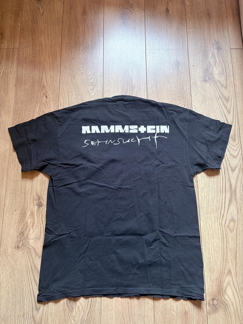 May include: Black t-shirt with the band name "RAMMSTEIN" in white block letters above the word "Sehnsucht" in a stylized white font. The shirt is laid flat on a wooden surface.