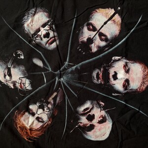 May include: Black t-shirt featuring a graphic design of six faces arranged in a circular pattern, with a shattered glass effect overlaying the image. The faces have a pale complexion and are in various states of distress.