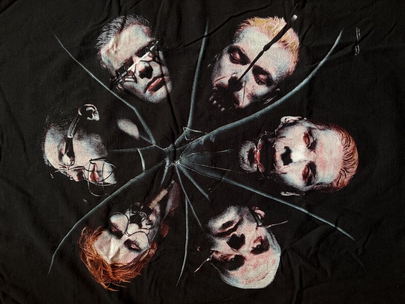 May include: Black t-shirt featuring a graphic design of six faces arranged in a circular pattern, with a shattered glass effect overlaying the image. The faces have a pale complexion and are in various states of distress.