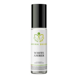 Puede incluir: Un frasco de vidrio transparente con aplicador de bola, tapa negra y etiqueta blanca. La etiqueta muestra un logotipo de mariposa verde, el texto AROMA SHORE y las palabras WHITE AMBER en negrita, seguidas de texto pequeño descriptivo.