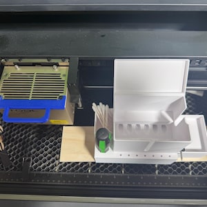 May include: A white, multi-compartment organizer with a hinged lid, next to a laser cutter head with a blue handle. A small wooden square and a green and black measuring tape are also visible.